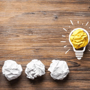 crumpled paper balls on a wooden table leading to a yellow paper shaped as a lightbulb, representing the shift from failed branding attempts to a strategic brand breakthrough for startups