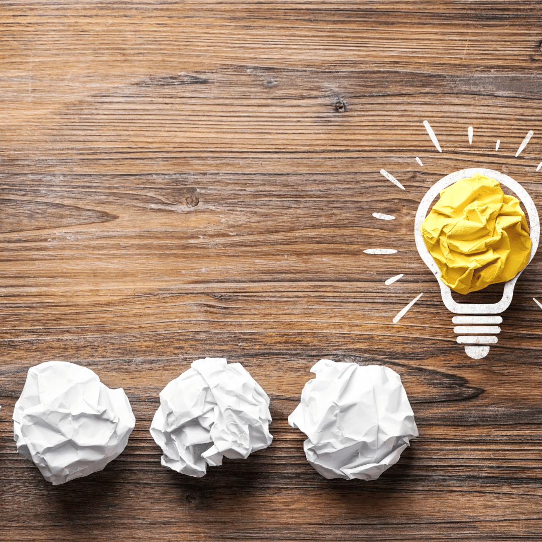crumpled paper balls on a wooden table leading to a yellow paper shaped as a lightbulb, representing the shift from failed branding attempts to a strategic brand breakthrough for startups