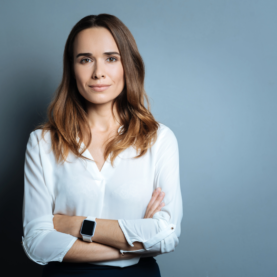 professional woman standing against a neutral blue background with arms crossed, wearing a white blouse and a smartwatch, conveying confidence and calm leadership.